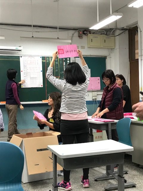 A woman holds up a paper while a man writes on a blackboard.