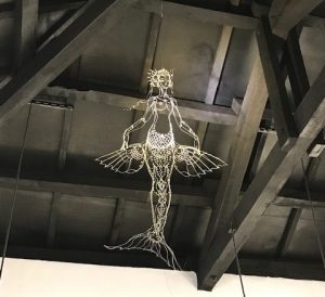 A mermaid sculpted out of wire hanging against ceiling rafters.