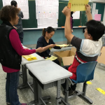 A man is holding up a ballot while another man records the vote on a blackboard. Two women are removing ballots from a box and handing them to the man calling out the result.