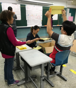A man is holding up a ballot while another man records the vote on a blackboard. Two women are removing ballots from a box and handing them to the man calling out the result.