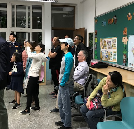 A group of people in a school classroom, all facing the same direction, watching something.